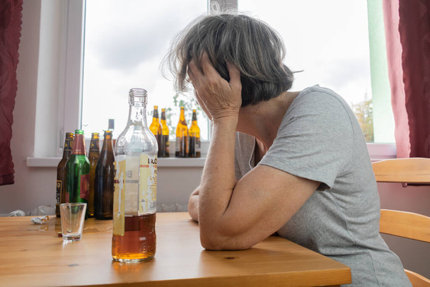 ηλικιωμένη ηλικιωμένη γυναίκα κάθεται δίπλα στο τραπέζι πίνουν αλκοόλ μπουκάλι στο σπίτι λυπημένος μόνος αλκοολισμός Σημεία και συμπτώματα κατάχρησης αποκατάστασης και προβλήματα αποκατάστασης - Φωτογραφία, εικόνα