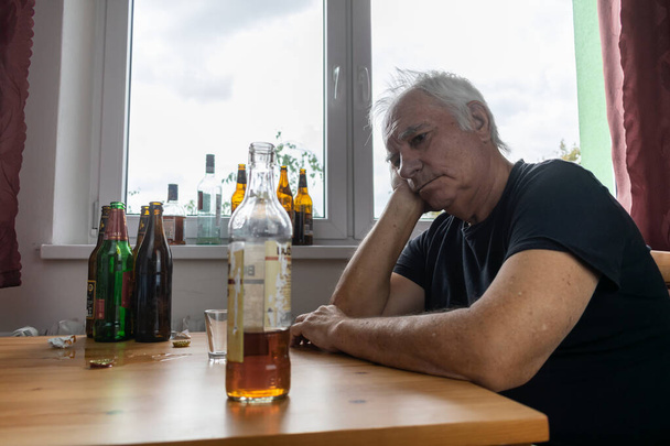 oude oudere man zitten naast tafel drinken alcohol fles thuis verdrietig alleen alcoholisme tekenen en symptomen afkicken misbruik en herstel problemen - Foto, afbeelding