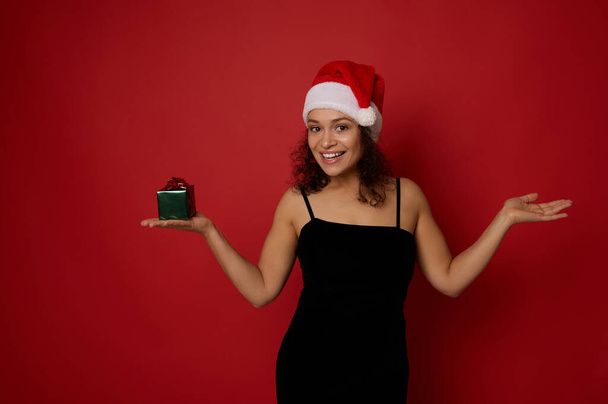 Mixed race pretty woman wearing a Santa hat, smiles toothy smile looking at camera, holds a Christmas gift in one hand and lifts the other hand palm up holding imaginary copy space on red background - Фото, зображення