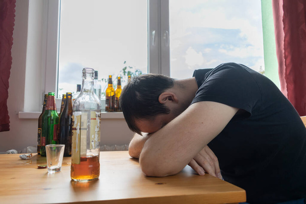 Молодой мужчина сидит рядом со столом пить бутылку алкоголя дома грустно одинокий алкоголизм Знаки и симптомы реабилитации злоупотребления и проблемы восстановления - Фото, изображение