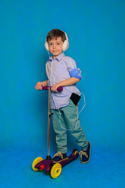 Un niño en una moto con auriculares sobre un fondo azul. Un artículo sobre scooters infantiles. Un artículo sobre la elección de un scooter para un niño. Un artículo sobre la seguridad del niño. Un niño feliz. Fondo azul y un niño. Copiar espacio - Foto, imagen