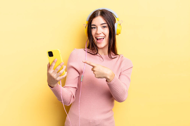 young hispanic woman smiling cheerfully, feeling happy and pointing to the side. headphones and telephone concept - Foto, Bild
