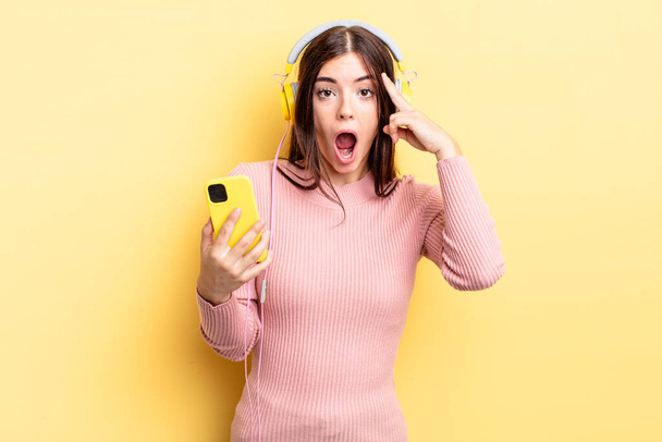 young hispanic woman looking surprised, realizing a new thought, idea or concept. headphones and telephone concept - Fotó, kép