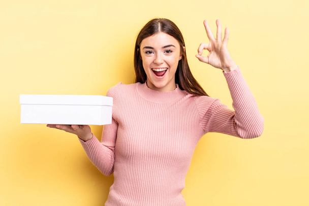 young hispanic woman feeling happy, showing approval with okay gesture. empty box concept - Foto, Imagen