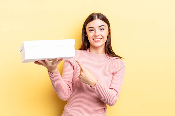 young hispanic woman smiling cheerfully, feeling happy and pointing to the side. empty box concept - Fotoğraf, Görsel