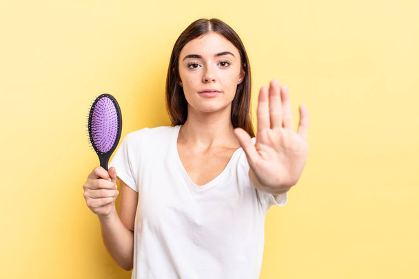 young hispanic woman looking serious showing open palm making stop gesture. hair brush concept - Foto, Imagen