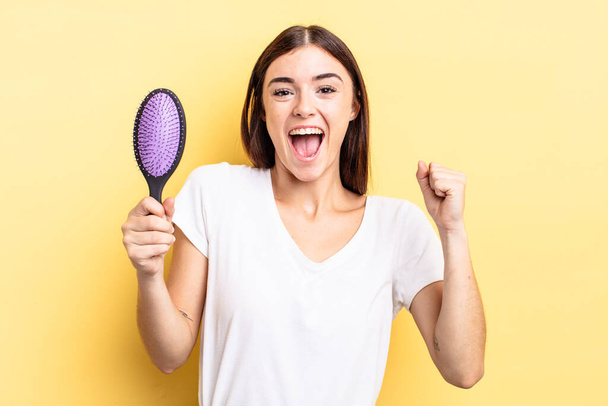 young hispanic woman feeling shocked,laughing and celebrating success. hair brush concept - Photo, Image