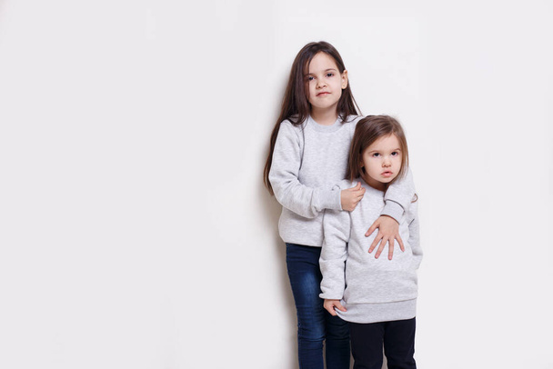 Δύο αξιολάτρευτο κοριτσάκι σε πουλόβερ και τζιν, ποζάρουν μαζί σε λευκό φόντο, χαμογελώντας και κοιτάζοντας την κάμερα. - Φωτογραφία, εικόνα