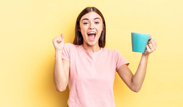 young hispanic woman feeling shocked,laughing and celebrating success. coffee cup concept - Photo, Image