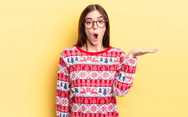 young hispanic woman looking surprised and shocked, with jaw dropped holding an object. chistmas and new year concept - Фото, изображение