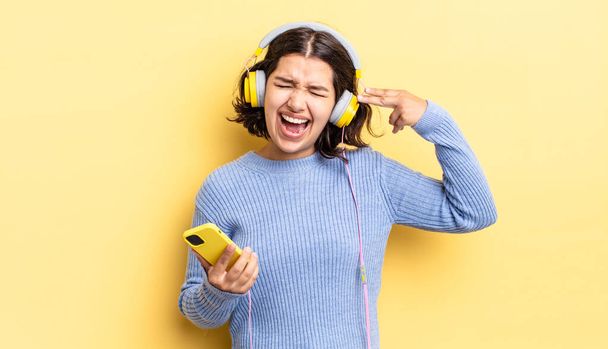 young hispanic woman looking unhappy and stressed, suicide gesture making gun sign. headphones and smartphone concept - 写真・画像