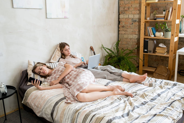 Deux filles reposantes utilisant des gadgets mobiles tout en étant couché sur le lit - Photo, image