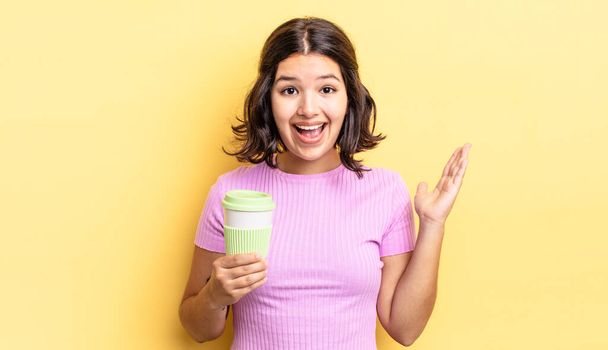 young hispanic woman feeling happy and astonished at something unbelievable. take away coffee concept - Фото, изображение