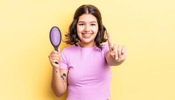 young hispanic woman smiling and looking friendly, showing number one. hairbrush concept - Foto, Imagem