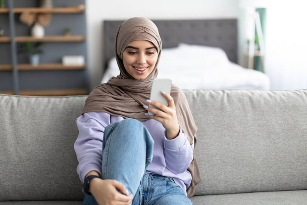 Χαμογελώντας islamic κυρία χρησιμοποιώντας το κινητό τηλέφωνο κάθεται στον καναπέ - Φωτογραφία, εικόνα