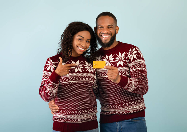 Concepto de compras de Navidad. Pareja negra cariñosa en suéteres cálidos de Navidad apuntando a la tarjeta de crédito, fondo azul - Foto, Imagen