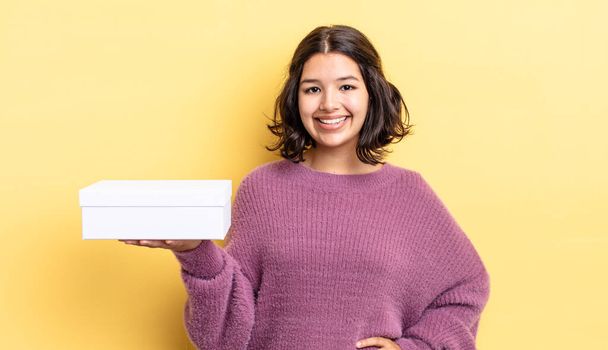 young hispanic woman smiling happily with a hand on hip and confident. blank box concept - Photo, Image