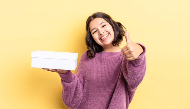 young hispanic woman feeling proud,smiling positively with thumbs up. blank box concept - Zdjęcie, obraz