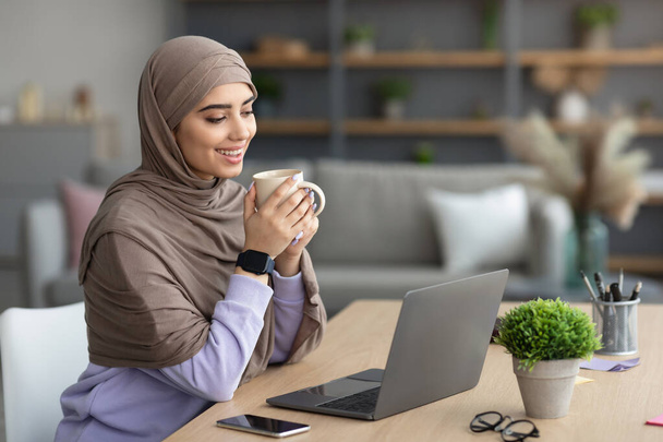 Muzułmanka ogląda wideo na komputerze, pije gorącą kawę - Zdjęcie, obraz