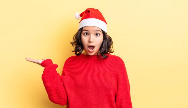 young hispanic woman looking surprised and shocked, with jaw dropped holding an object. christmas concept - Фото, изображение