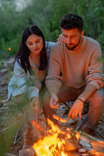 Молодая любящая пара жарит сосиски над огнем - Фото, изображение