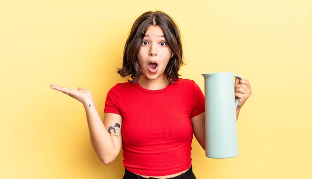 young hispanic woman looking surprised and shocked, with jaw dropped holding an object. thermos concept - Photo, Image