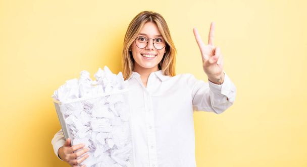 pretty blonde girl smiling and looking happy, gesturing victory or peace. paper balls trash concept - Photo, Image