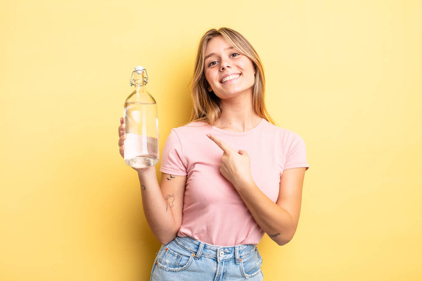 pretty blonde girl smiling cheerfully, feeling happy and pointing to the side. water bottle concept - Fotó, kép