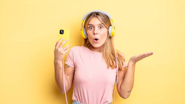 pretty blonde girl looking surprised and shocked, with jaw dropped holding an object. listening music concept - Foto, imagen