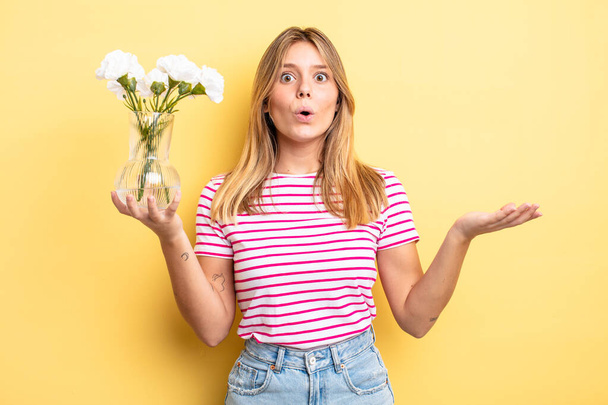 pretty blonde girl looking surprised and shocked, with jaw dropped holding an object. decorative flowers concept - Foto, afbeelding