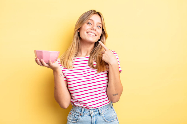 pretty blonde girl smiling confidently pointing to own broad smile. empty bowl concept - Foto, Imagen