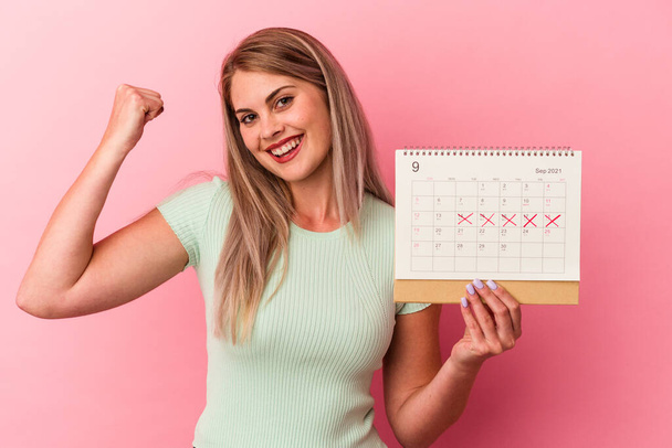 Joven mujer caucásica sosteniendo un calendario aislado sobre fondo rosa levantando puño después de una victoria, concepto ganador. - Foto, Imagen