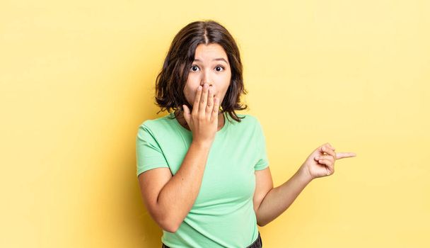 young pretty girl feeling happy, shocked and surprised, covering mouth with hand and pointing to lateral copy space - Photo, Image