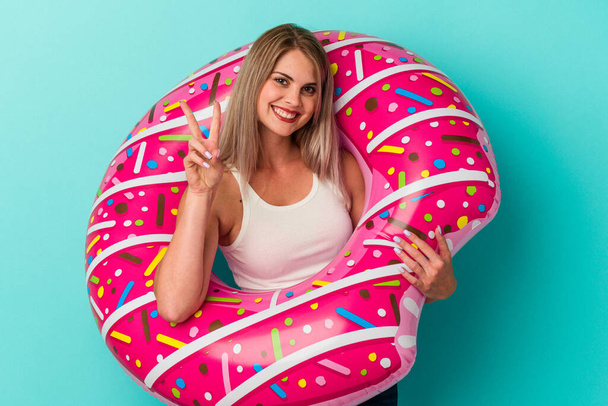 jonge russische vrouw met opblaasbare donut geïsoleerd op blauwe achtergrond tonen nummer twee met vingers. - Foto, afbeelding