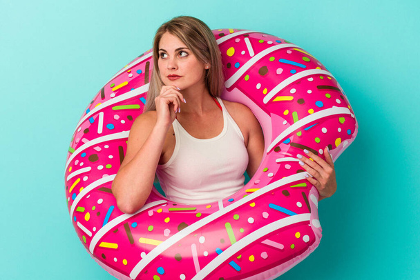 Young russian woman with inflatable donut isolated on blue background looking sideways with doubtful and skeptical expression. - Photo, Image