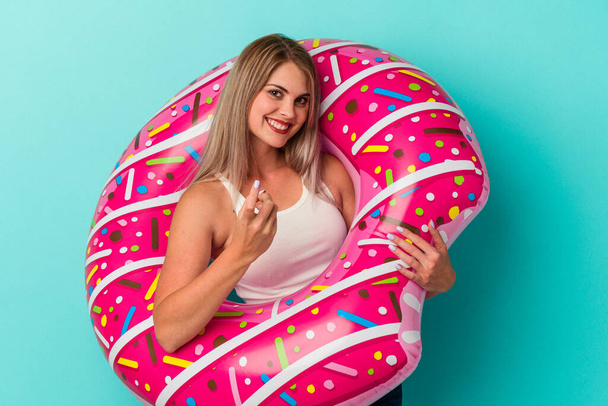 jonge russische vrouw met opblaasbare donut geïsoleerd op blauwe achtergrond wijzend met de vinger naar je alsof uitnodigen dichterbij komen. - Foto, afbeelding