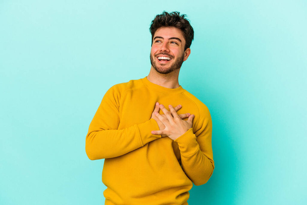 Молодий кавказький чоловік ізольований на синьому фоні сміється тримаючи руки на серці, концепція щастя
. - Фото, зображення