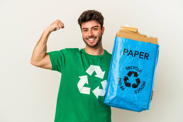 Hombre joven caucásico reciclando cartón aislado sobre fondo blanco levantando puño después de una victoria, concepto ganador. - Foto, Imagen