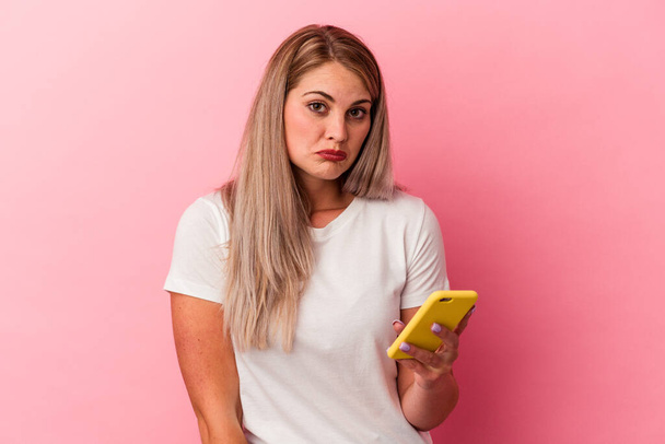 Young russian woman holding a mobile phone isolated on pink background shrugs shoulders and open eyes confused. - Foto, Bild