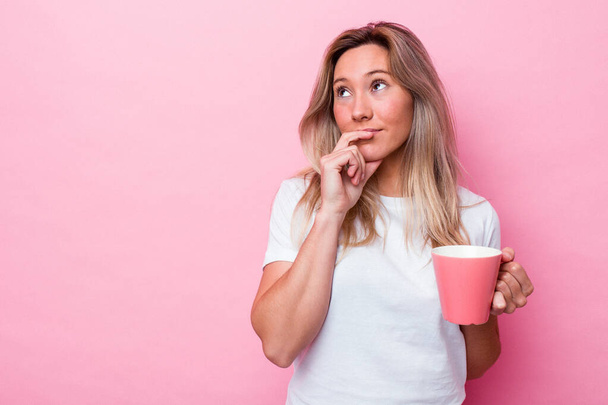 Giovane donna australiana che tiene una tazza rosa isolata su sfondo rosa guardando lateralmente con espressione dubbia e scettica. - Foto, immagini