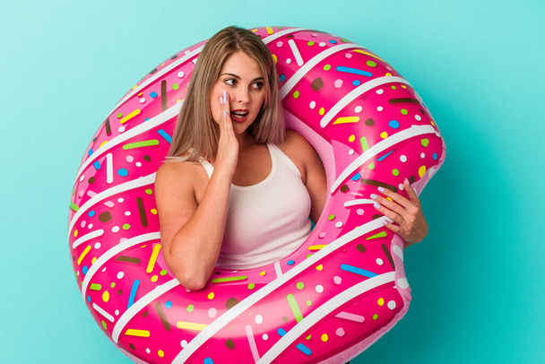 Young russian woman with inflatable donut isolated on blue background is saying a secret hot braking news and looking aside - Photo, Image