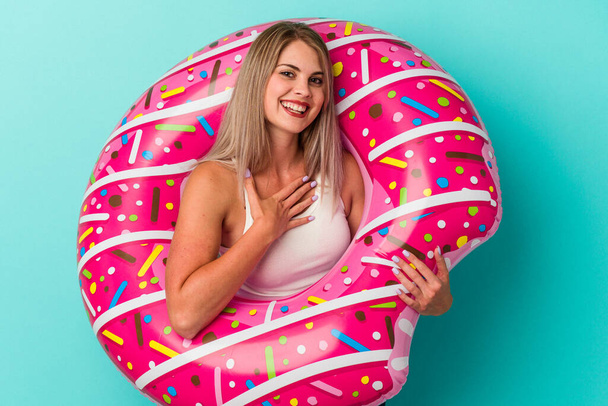 Young russian woman with inflatable donut isolated on blue background laughs out loudly keeping hand on chest. - 写真・画像