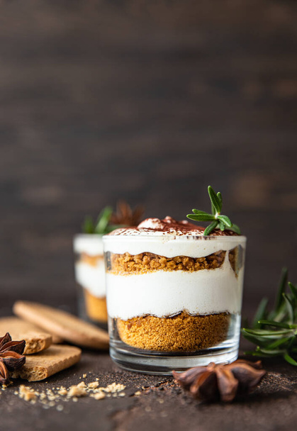 Layered dessert in glass jar with cookie crumble and whipped cream decorated with rosemary and anise, dark brown background. No bake cheesecake, trifle or pudding. - Zdjęcie, obraz