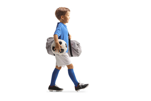 Full length profile shot of a boy wearing a sports jersey, holding a soccer ball and bag and walking isolated on white background - Zdjęcie, obraz