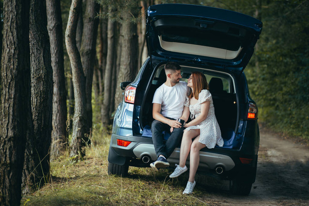 Young couple, man and woman, hugging together on a picnic, sitting in the trunk of a car in the woods, happy together - Фото, зображення