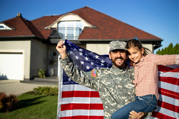 Portret ojca żołnierza w mundurze na przepustce wojskowej trzymającego przed domem swoją uroczą córkę i amerykańską flagę.. - Zdjęcie, obraz