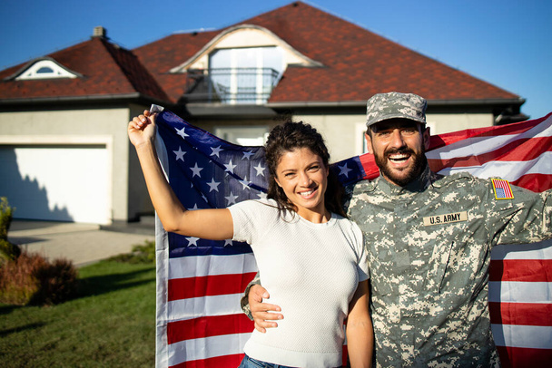 軍の休暇に制服を着た夫の兵士の肖像彼の美しい妻を抱きかかえ、彼らの家の前でアメリカの旗を保持. - 写真・画像