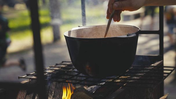 mélanger la nourriture dans une casserole avec la main, cuire la nourriture sur le feu dans un voyage de camping dans la nature, recettes cuites sur le feu et la fumée, activités de plein air modernes le week-end, la vapeur chaude monte d'une casserole - Photo, image