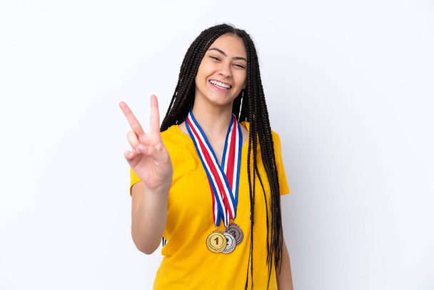 Teenager dívka s copánky a medaile přes izolované růžové pozadí s úsměvem a ukazující vítězství znamení - Fotografie, Obrázek