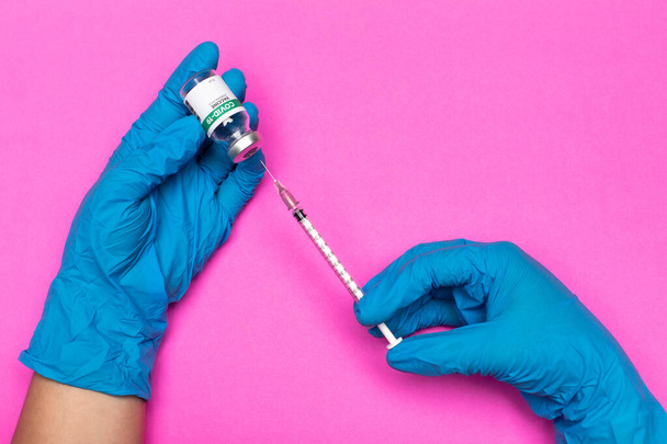 Pinkfarbener Hintergrund, medizinische Fachkräfte mit blauen Hygienehandschuhen, die Spritzennadel in versiegelte Glaskapsel injizieren, um Covid-19-Impfstoff für den Aktivierungstest der Krankheitsimmunität im Krankenhauslabor auszusaugen - Foto, Bild